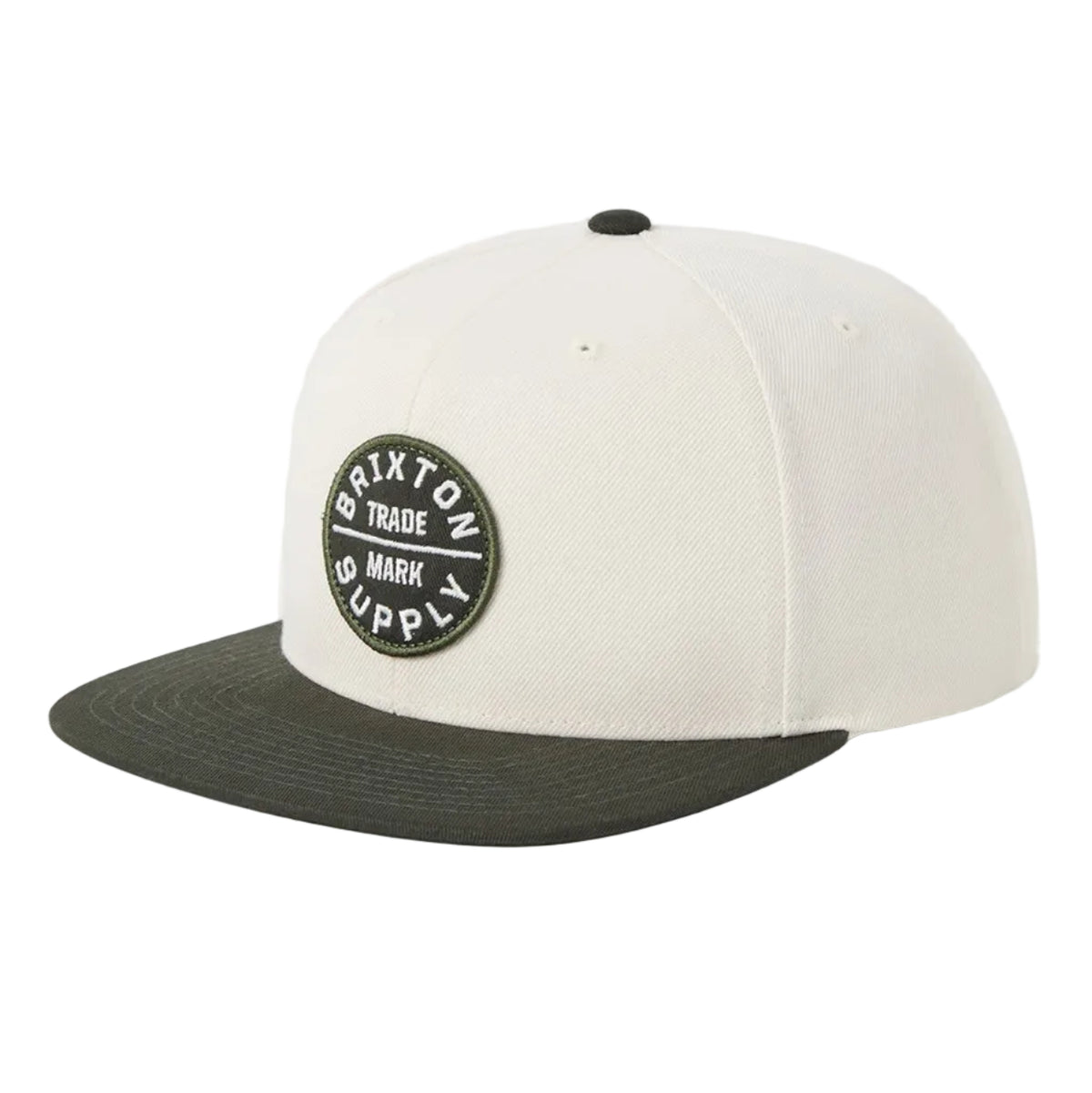 A structured baseball cap with an off-white crown and a dark olive green flat brim, featuring a circular black and white embroidered patch on the front that reads 'Brixton Trade Mark Supply'.