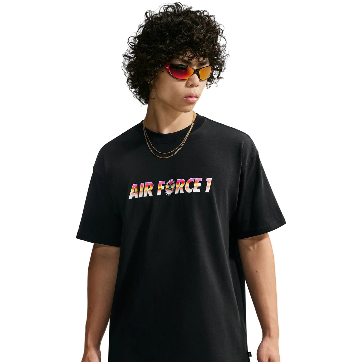 A model with curly black hair wearing a black t-shirt with 'AIR FORCE 1' printed across the chest in a colorful gradient pattern, styled with gold necklaces and red reflective sunglasses against a light gray background.