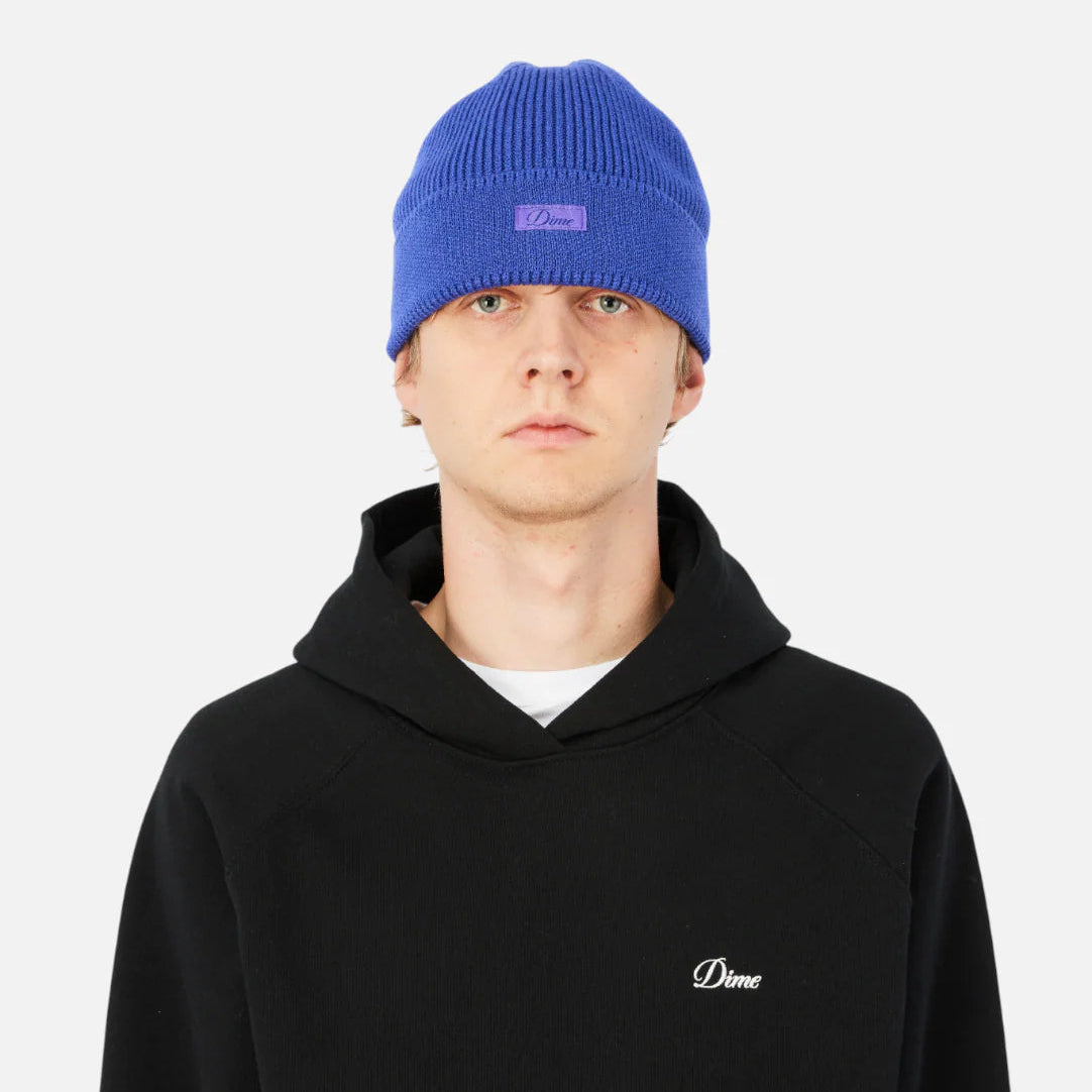 Person wearing a black hoodie with a small logo and a blue beanie on a white background