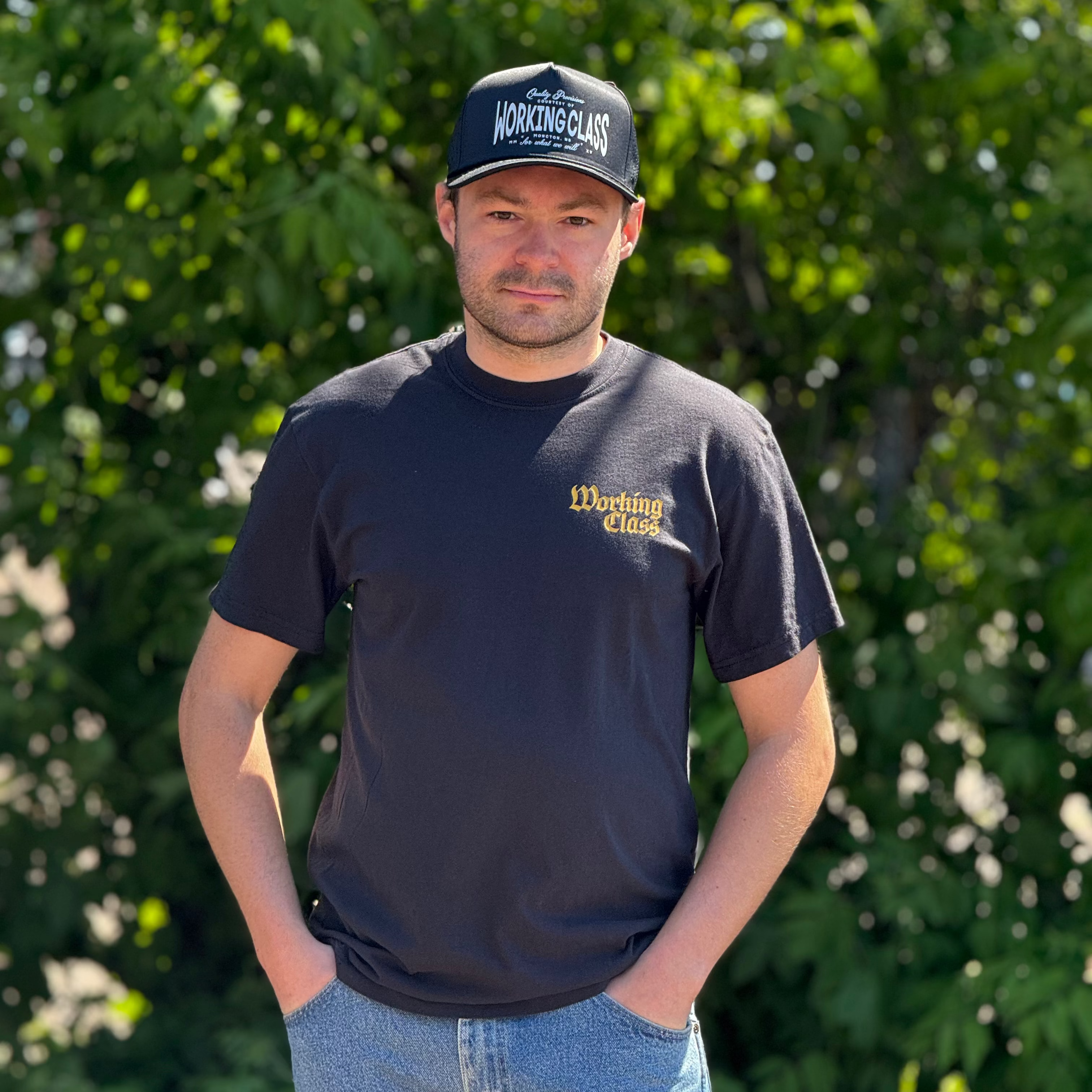A man wearing a black crewneck t-shirt with a gold 'Working Class' logo on the left chest, paired with a black baseball cap that reads 'WORKING CLASS' and blue jeans, standing outdoors in front of a green leafy background.