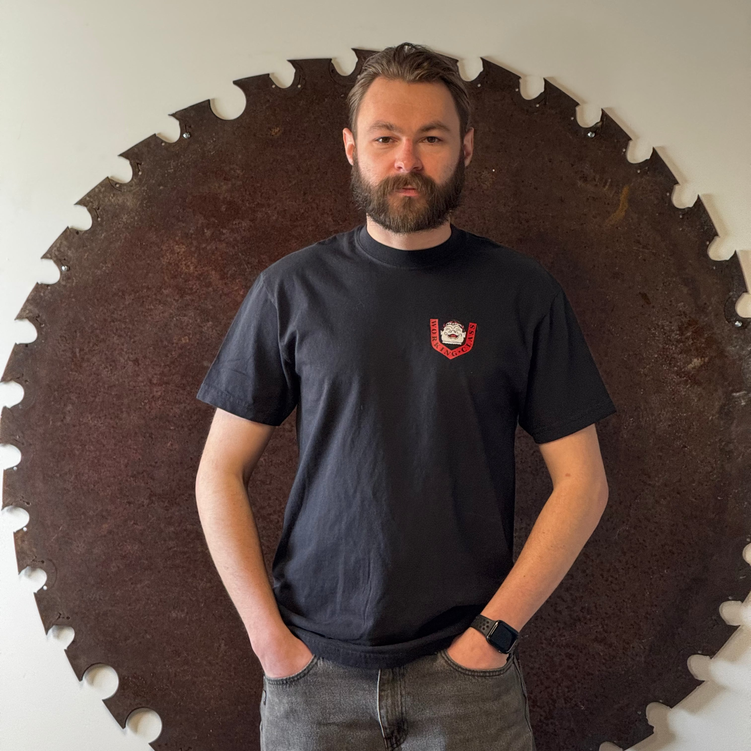 Man wearing a black t-shirt with a logo standing in front of a large circular saw blade.