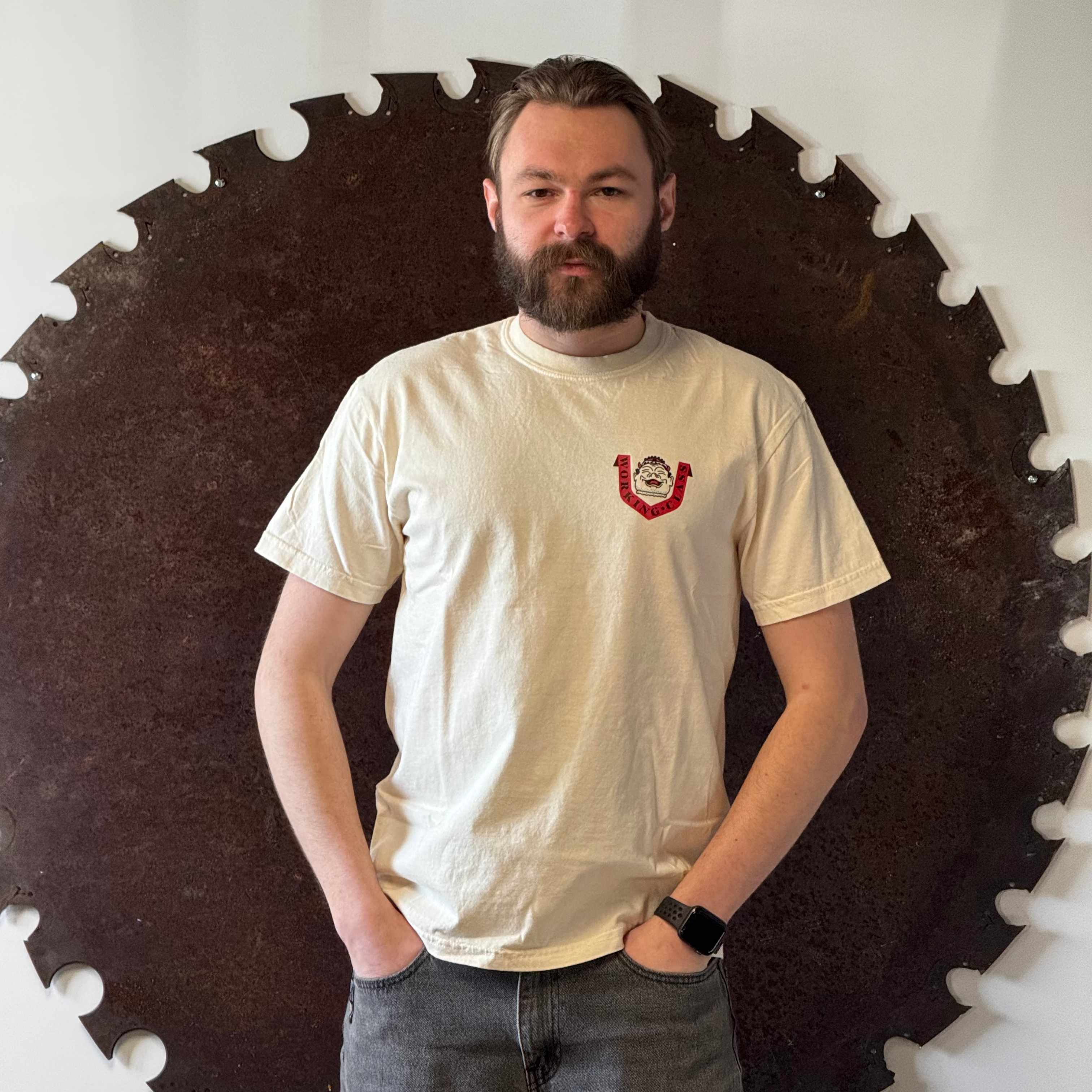 Man wearing a white t-shirt with a red logo standing in front of a large circular saw blade.