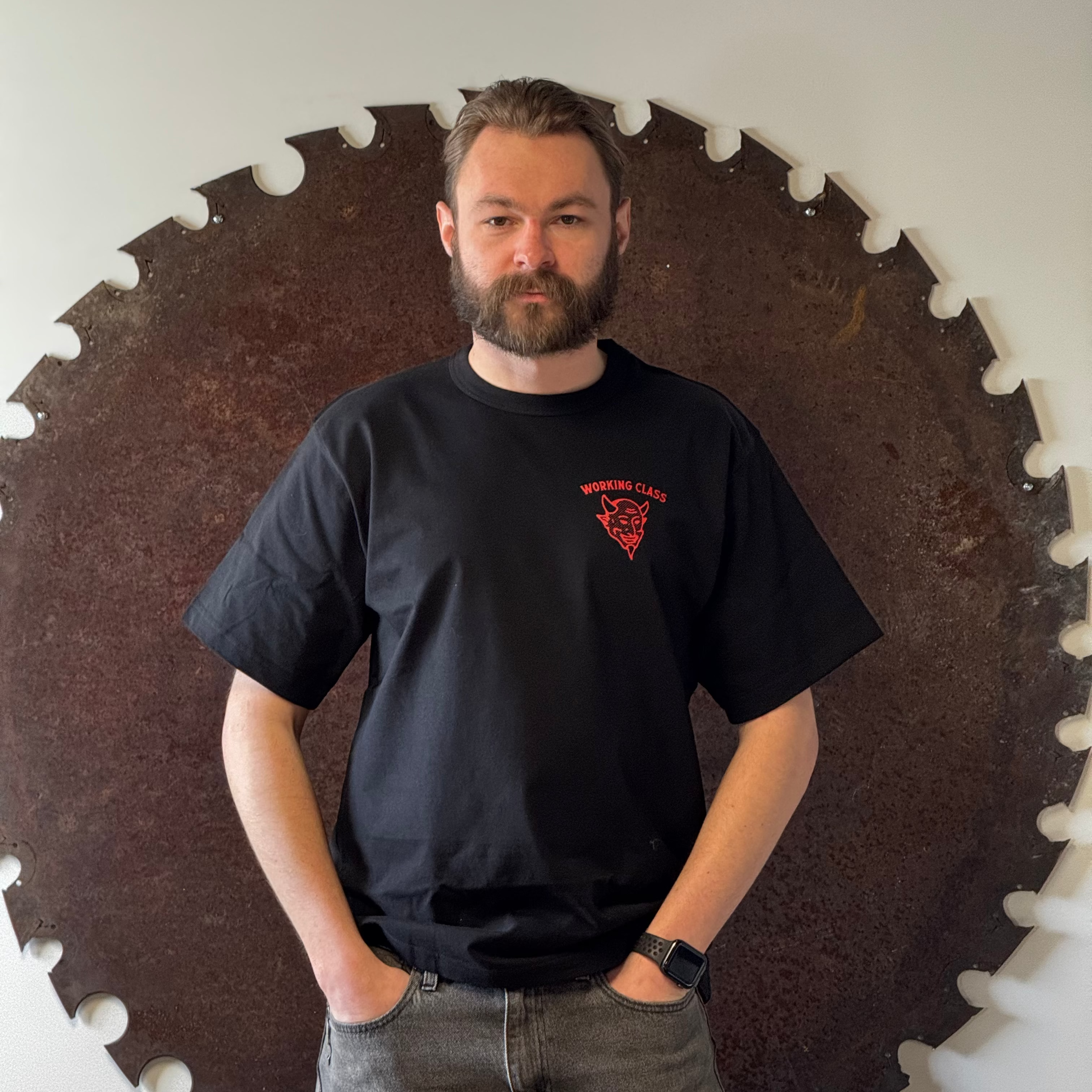 Man wearing a black t-shirt with a red logo standing in front of a large circular saw blade.