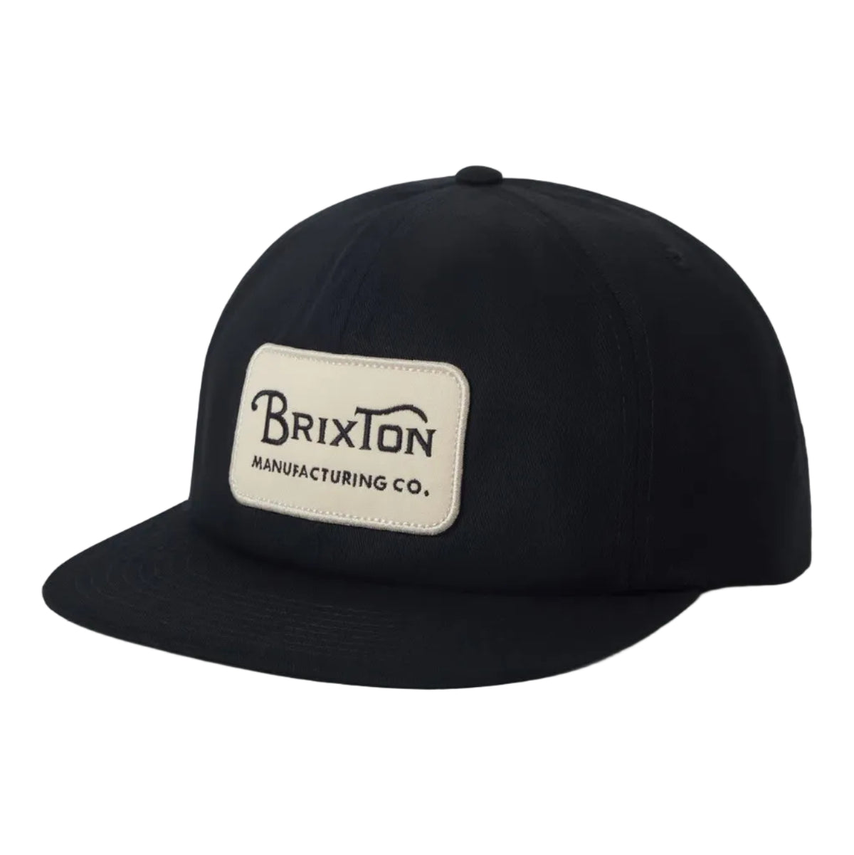 A black flat-brim baseball cap with a beige rectangular patch on the front that reads 'Brixton Manufacturing Co.' in black text, photographed on a clean light gray background.