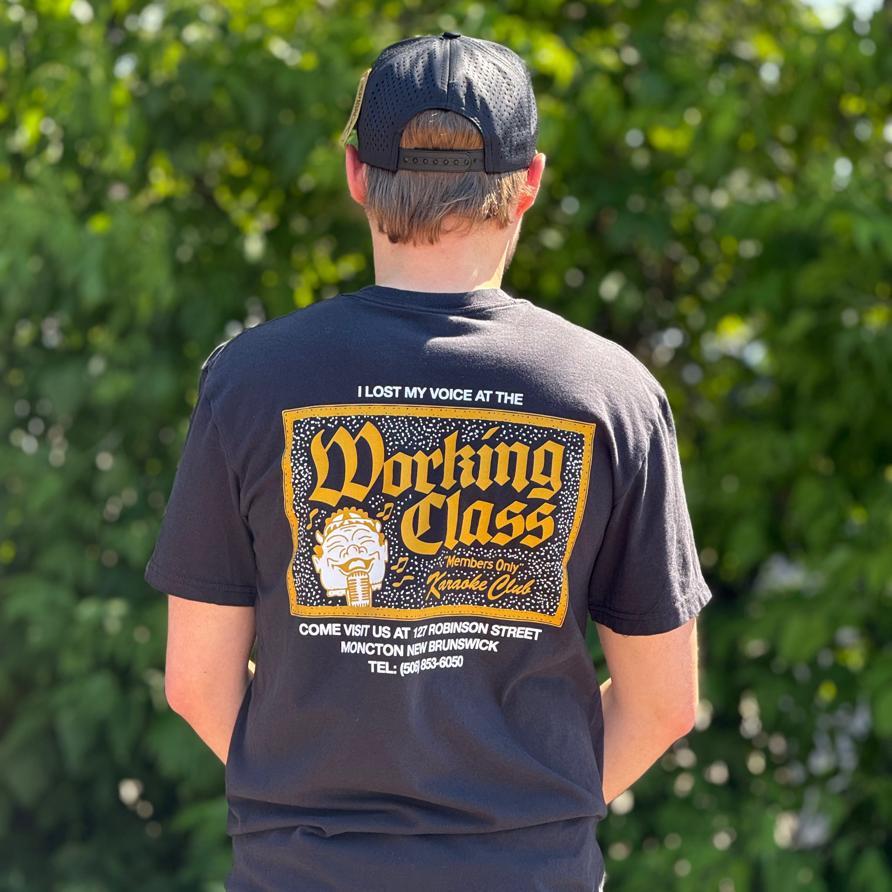 A man viewed from behind wearing a black graphic t-shirt with a yellow and white 'Working Class' design, paired with a black baseball cap and blue jeans, standing outdoors in front of a green leafy background.