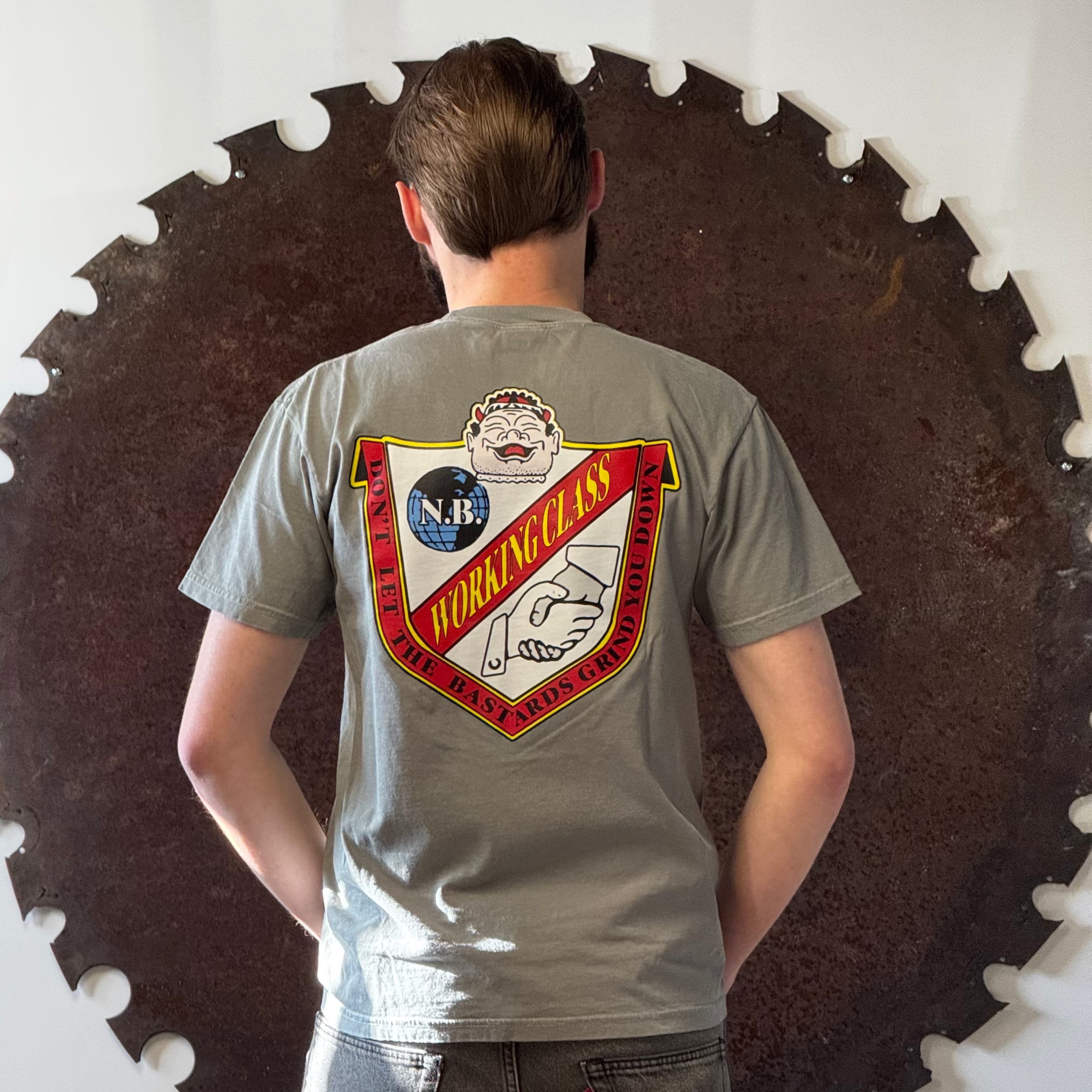 Person wearing a gray t-shirt with a logo on the back, standing in front of a large circular saw blade.
