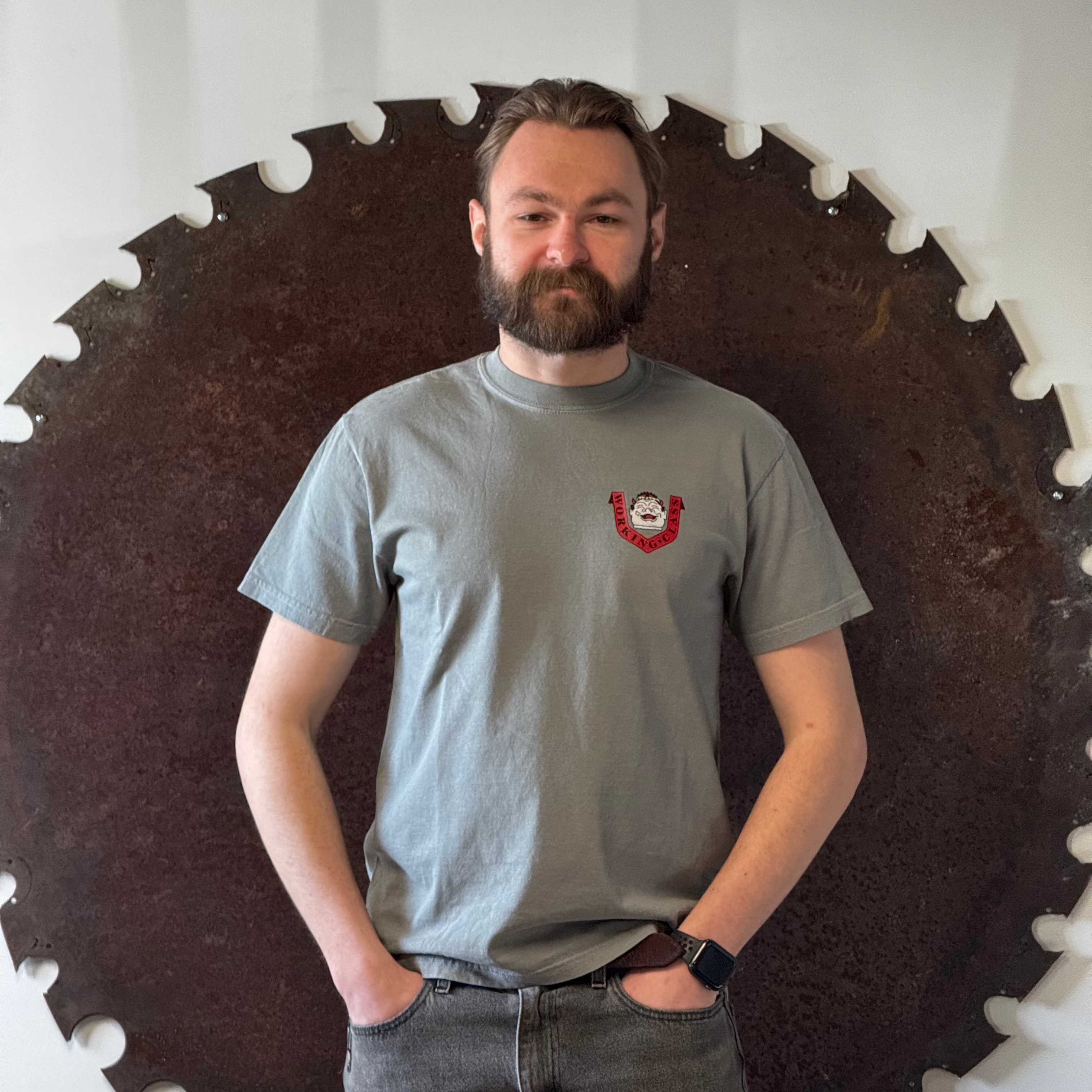 Man wearing a gray t-shirt with a logo standing in front of a large circular saw blade.