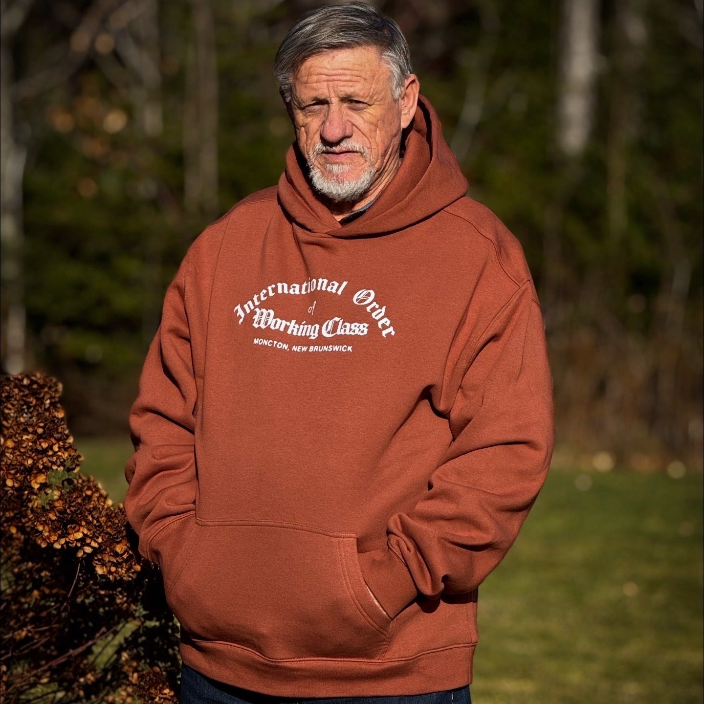 Man wearing a brown hoodie with text standing outdoors in a forest setting