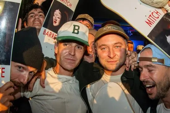 A group of men posing closely together in a crowded indoor setting, with posters and a skateboard visible in the background. The central figure wears a white cap with a green brim and a white shirt, while another man to his right wears a brown cap.