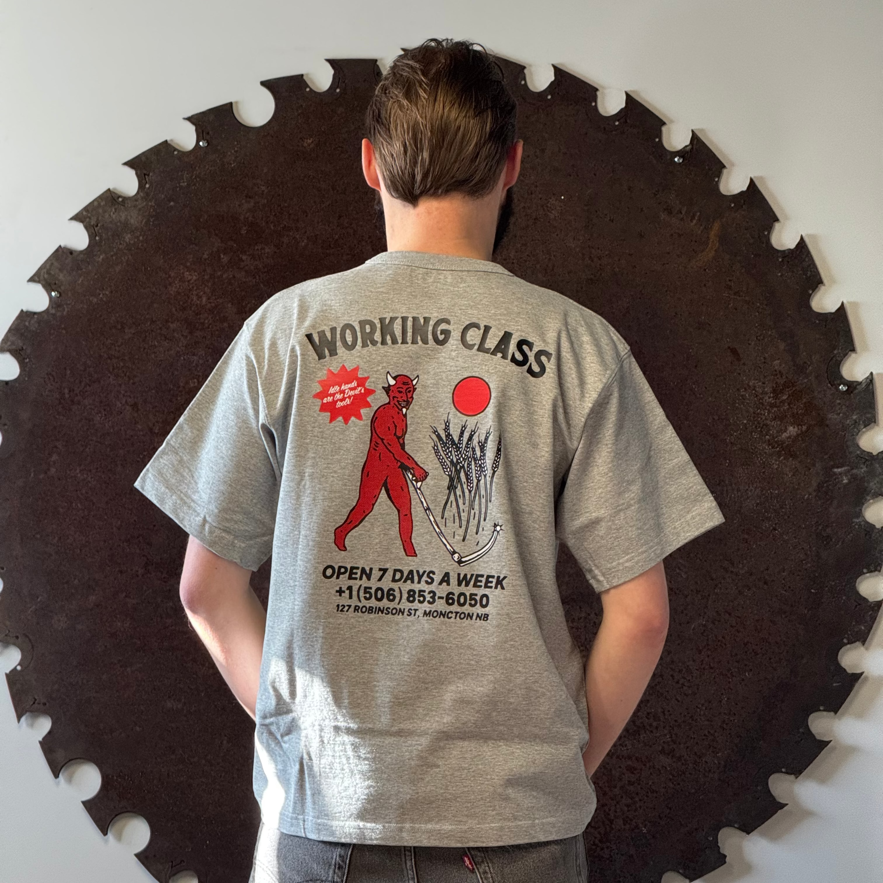 Person wearing a gray t-shirt with a graphic design and text, standing in front of a large circular saw blade.