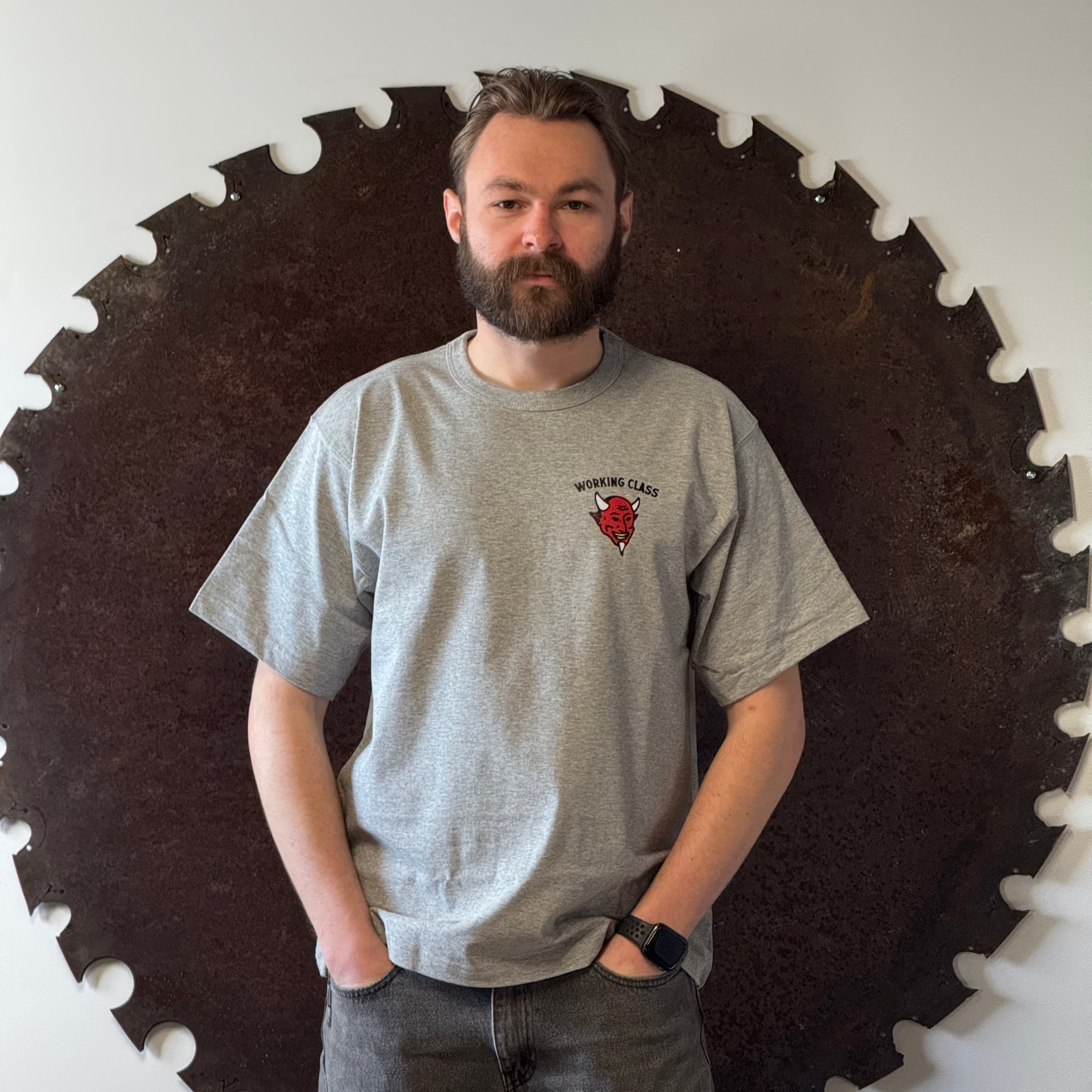 Man wearing a gray t-shirt with a logo standing in front of a large circular saw blade.