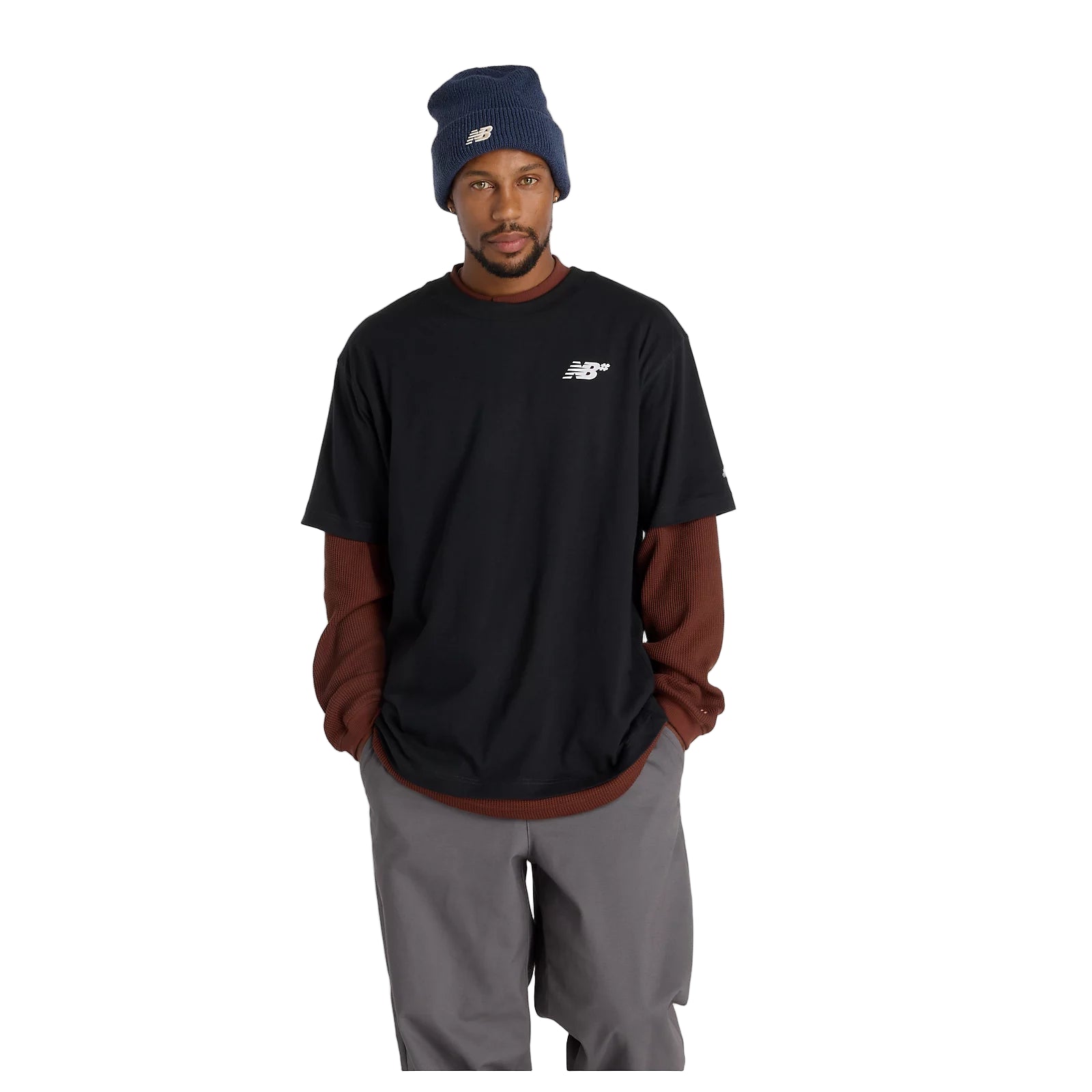 Man wearing a black t-shirt with a logo, brown long-sleeve shirt underneath, gray pants, and a navy beanie on a light gray background.