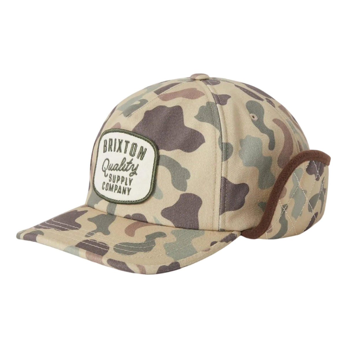 A camouflage trucker-style cap with ear flaps and a sewn-on patch reading 'Brixton Quality Supply Company', shown on a light gray background.